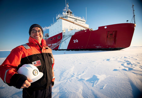 A Coast Guardsman stands on ice with an ice cutter in the background.
