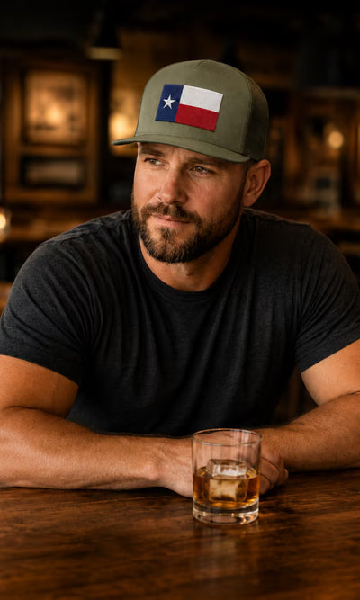 TXI Lids - man wearing a Texas flag trucker cap and black tee, seated at a bar.