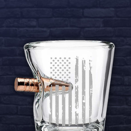Clear glass shot glass with American flag engraving and a bullet fused into it on the side, on a dark brick wall background.